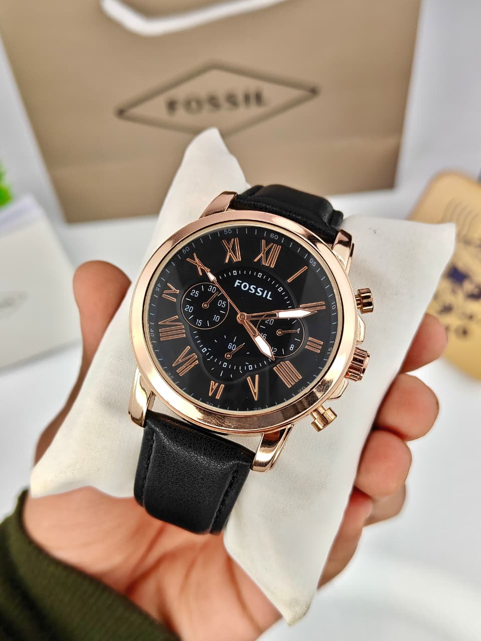 A Fossil chronograph watch with a black dial, black leather strap, and rose gold case, held in a hand over a white cushion with a Fossil shopping bag in the background.