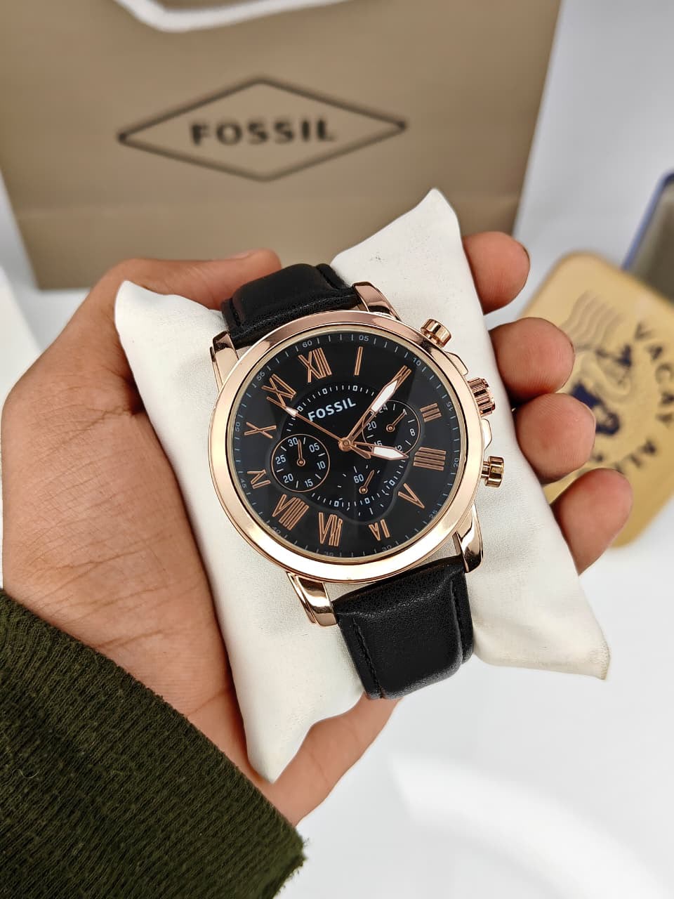 A Fossil wristwatch with a black dial, black leather strap, and rose gold case, held in a hand on a white cushion with the Fossil box in the background.