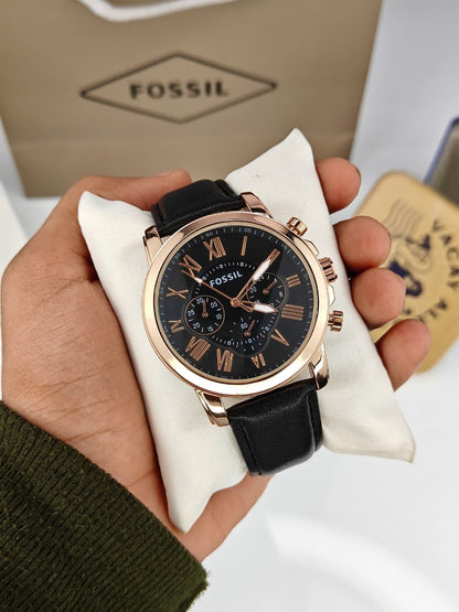 A Fossil wristwatch with a black dial, black leather strap, and rose gold case, held in a hand on a white cushion with the Fossil box in the background.
