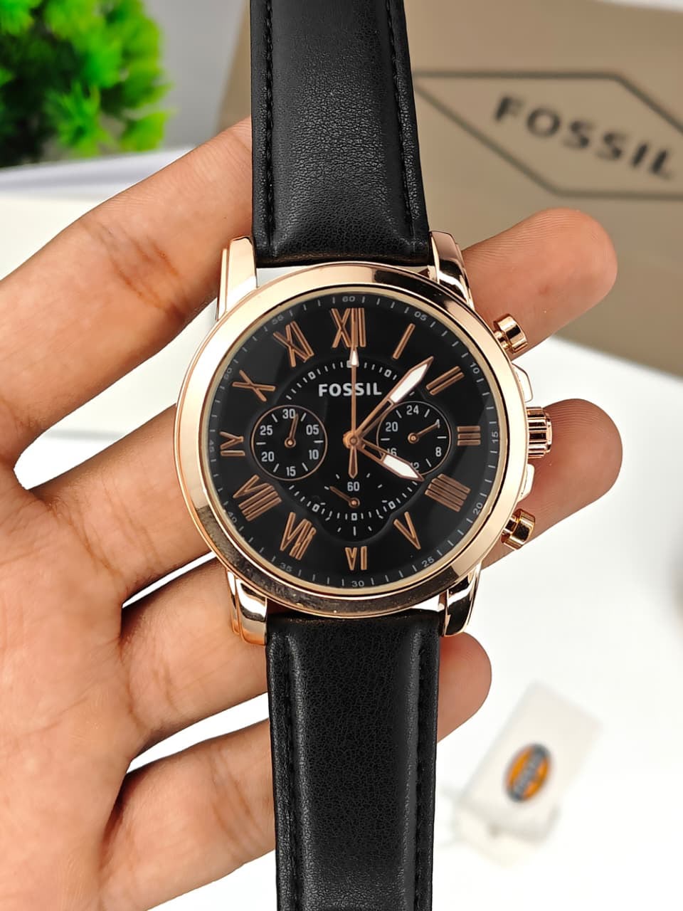 A Fossil chronograph watch with a black dial, black leather strap, and a rose gold-tone case, held in a person's hand.