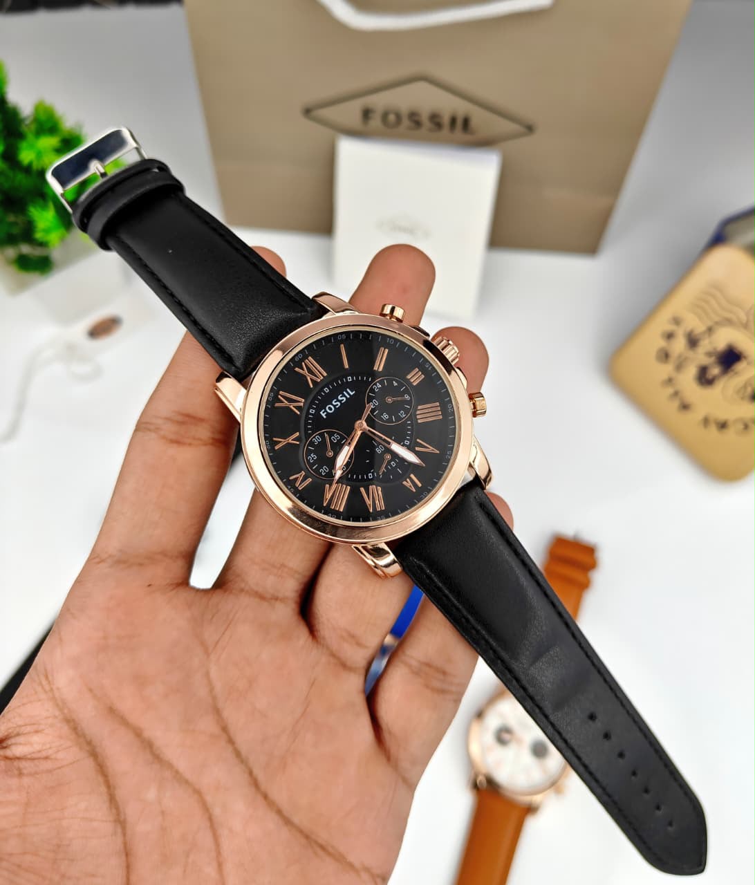 A Fossil chronograph watch with a black dial and black leather strap, held in a person's hand. The watch features a rose gold-toned case and matching rose gold hands and markers.